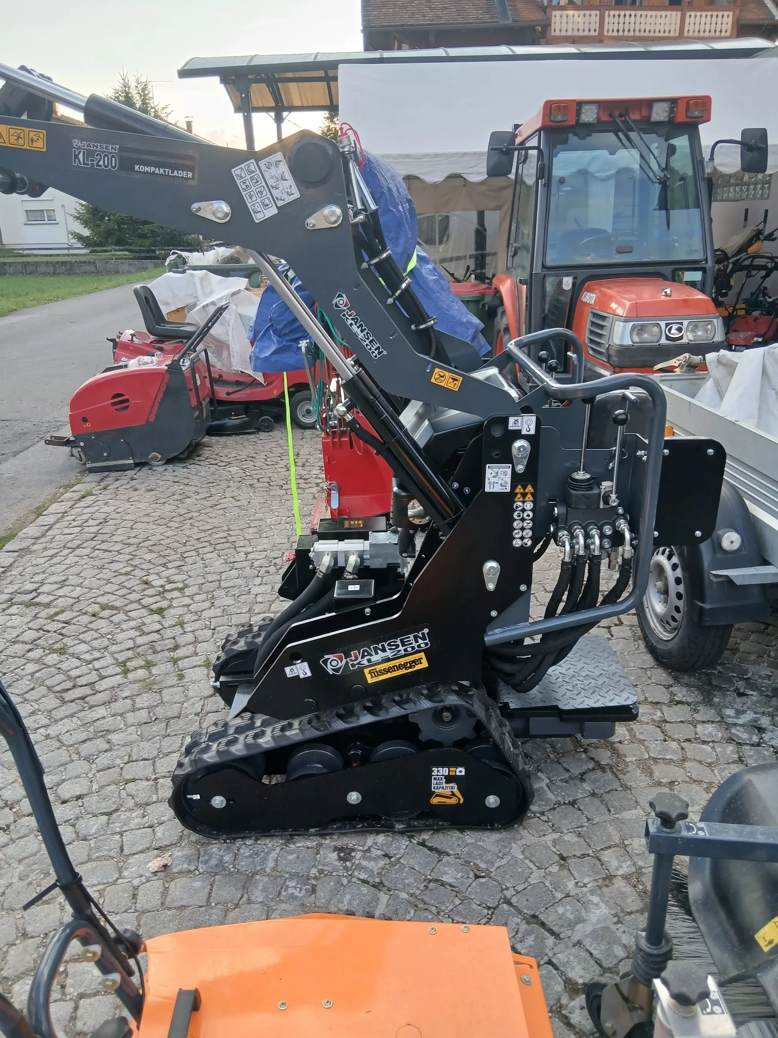Kompaktlader mit Fussenegger Aufkleber im Hof, dahinter Traktor und Anhänger