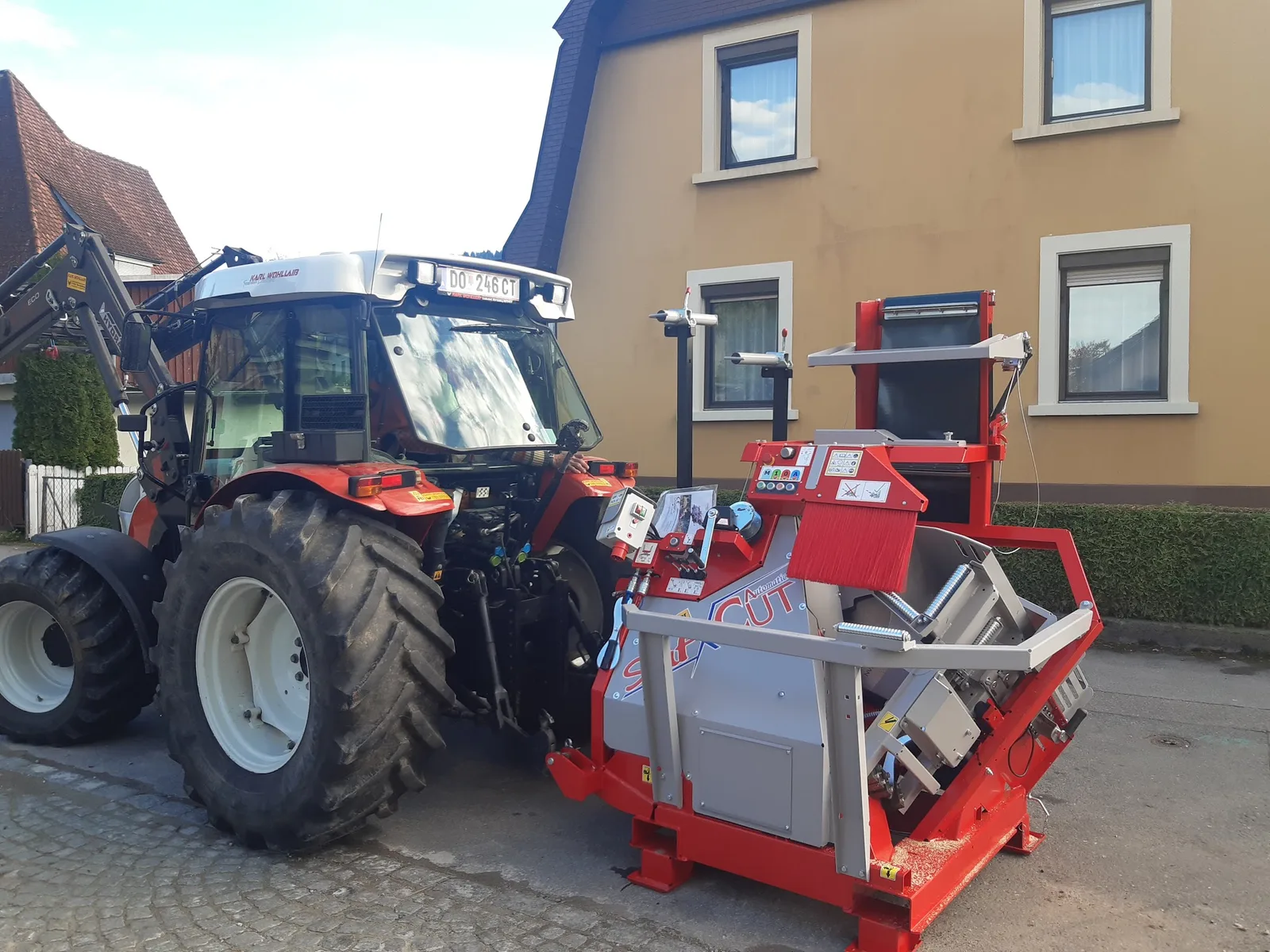 Traktor mit angebautem roten Sägeautomaten vor einem Haus