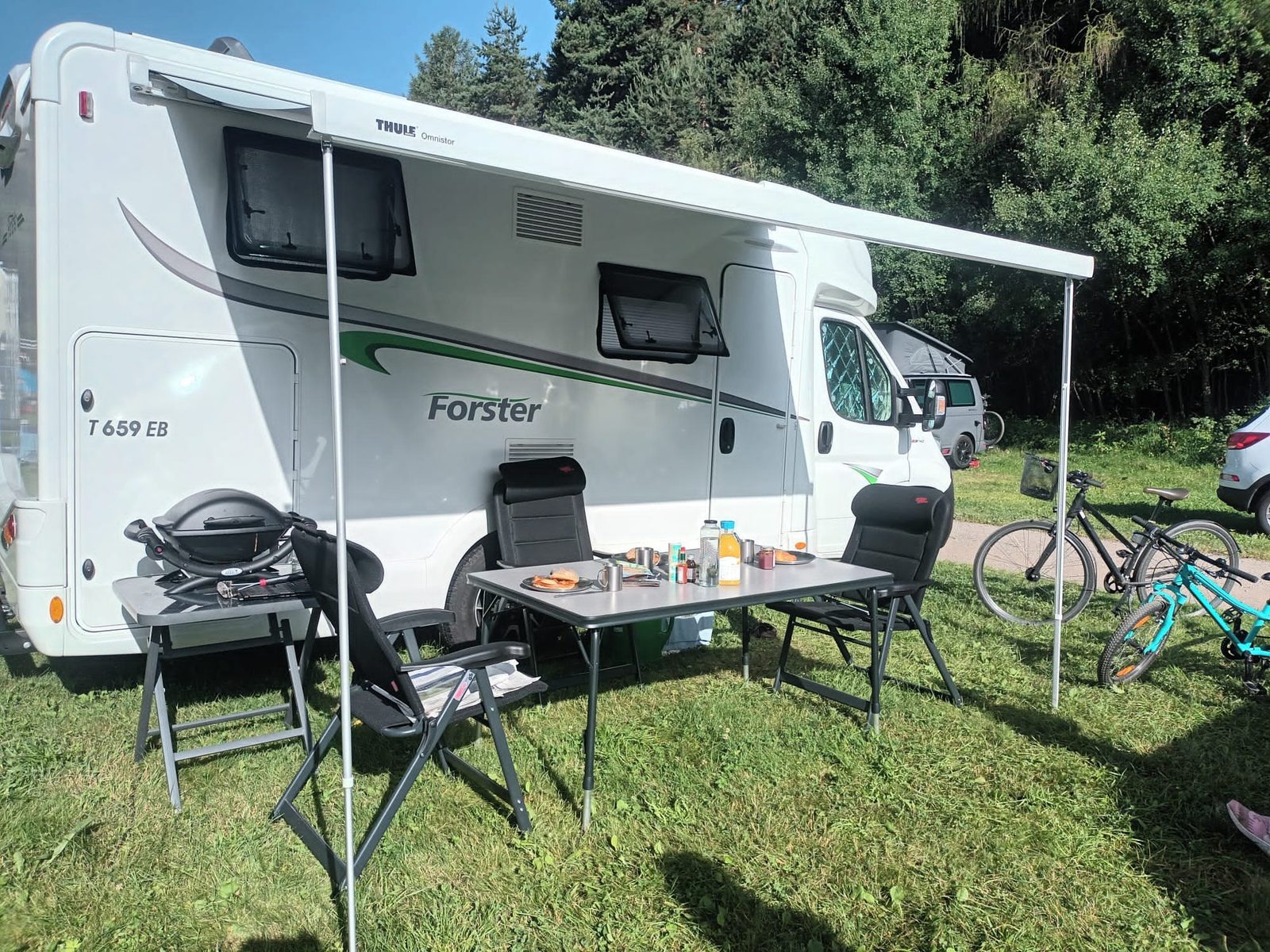 Wohnmobil mit ausgefahrener Markise, davor Tisch und Campingstühle auf einer Wiese
